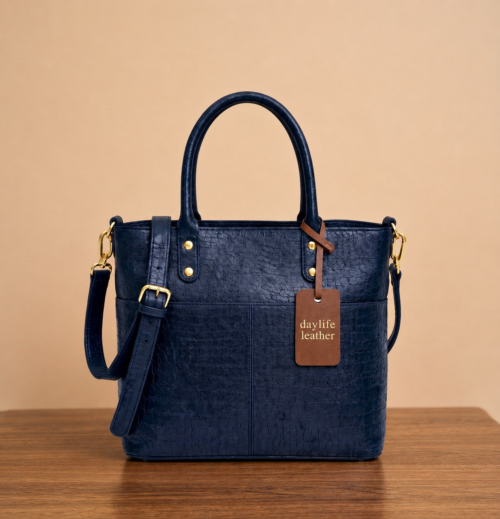 Front view of a dark blue croc-embossed leather tote bag by DayLife Leather, featuring gold hardware, an adjustable strap, and a brown leather logo tag, resting on a wooden table.