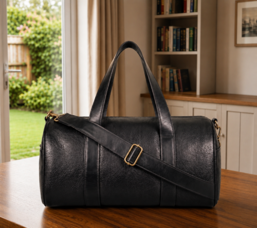 Premium black genuine leather duffle bag with adjustable shoulder strap, displayed in a modern lifestyle setting with natural lighting.