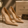 Nude beige premium leather pointed-toe high heel pumps with a gold buckle by DayLife Leather, placed next to luxury packaging box.