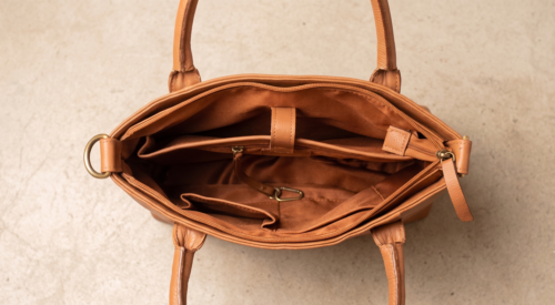 Open profile view of a DayLife Leather women's shoulder bag in a peach-pink hue, resting on a dark wood table with blurred background books.
