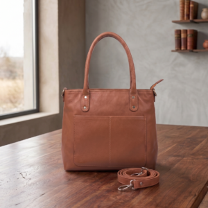 Side profile view of a DayLife Leather women's shoulder bag in a peach-pink hue, resting on a dark wood table with blurred background books.
