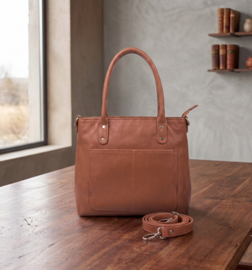 Side profile view of a DayLife Leather women's shoulder bag in a peach-pink hue, resting on a dark wood table with blurred background books.