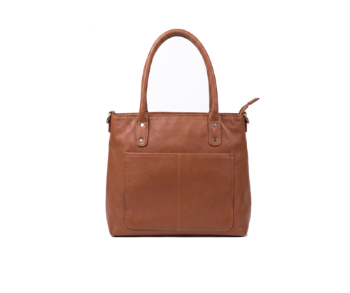 Side profile view of a DayLife Leather women's shoulder bag in a peach-pink hue, resting on a dark wood table with blurred background books.