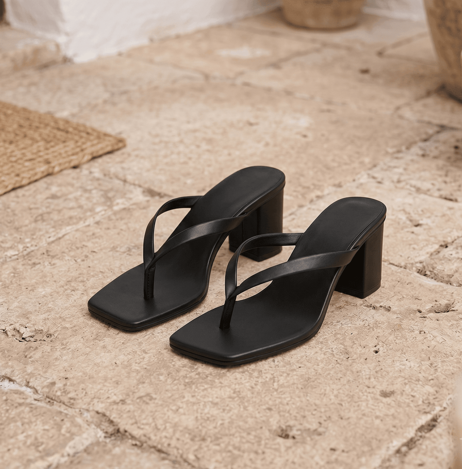 A pair of DayLife Leather black square-toe heeled sandals with thong-style straps, displayed on a rustic stone tiled floor surrounded by terracotta planters at DayLifeLeather.com.