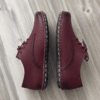 Close-up of feet wearing stylish maroon leather lace-up shoes with white contrast stitching, cuffed blue jeans, and a blurred home interior background.