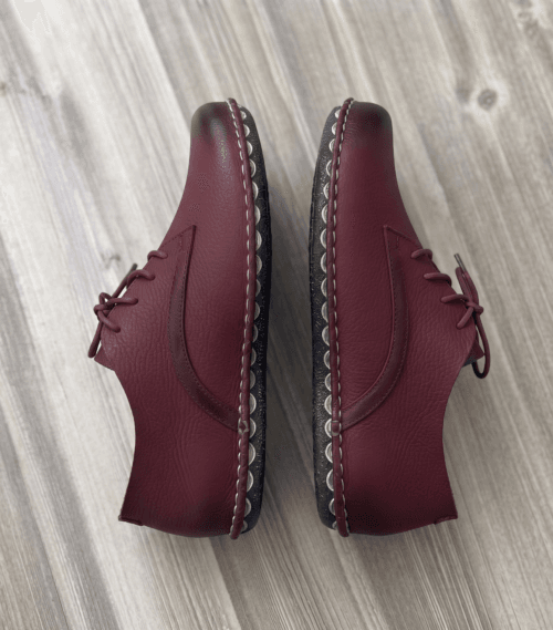 Close-up of feet wearing stylish maroon leather lace-up shoes with white contrast stitching, cuffed blue jeans, and a blurred home interior background.