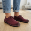 Close-up of feet wearing stylish maroon leather lace-up shoes with white contrast stitching, cuffed blue jeans, and a blurred home interior background.