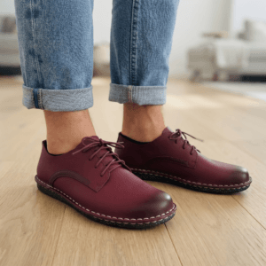 Handcrafted Maroon Leather Shoes with Contrast Stitching - Lifestyle View Close-up of feet wearing stylish maroon leather lace-up shoes with white contrast stitching, cuffed blue jeans, and a blurred home interior background.