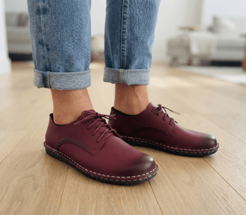 Close-up of feet wearing stylish maroon leather lace-up shoes with white contrast stitching, cuffed blue jeans, and a blurred home interior background.