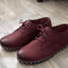 Close-up of feet wearing stylish maroon leather lace-up shoes with white contrast stitching, cuffed blue jeans, and a blurred home interior background.