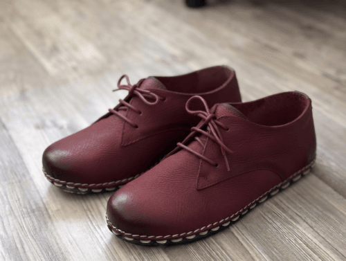 Close-up of feet wearing stylish maroon leather lace-up shoes with white contrast stitching, cuffed blue jeans, and a blurred home interior background.