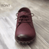 Close-up of feet wearing stylish maroon leather lace-up shoes with white contrast stitching, cuffed blue jeans, and a blurred home interior background.
