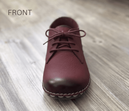 Close-up of feet wearing stylish maroon leather lace-up shoes with white contrast stitching, cuffed blue jeans, and a blurred home interior background.