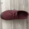 Close-up of feet wearing stylish maroon leather lace-up shoes with white contrast stitching, cuffed blue jeans, and a blurred home interior background.