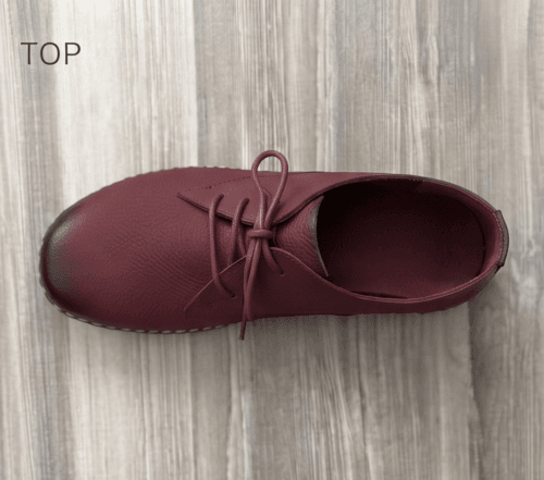 Close-up of feet wearing stylish maroon leather lace-up shoes with white contrast stitching, cuffed blue jeans, and a blurred home interior background.