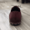 Close-up of feet wearing stylish maroon leather lace-up shoes with white contrast stitching, cuffed blue jeans, and a blurred home interior background.