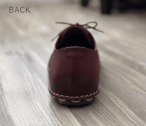 Close-up of feet wearing stylish maroon leather lace-up shoes with white contrast stitching, cuffed blue jeans, and a blurred home interior background.
