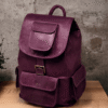 n aged purple leather backpack with multiple pockets on a wooden surface in front of a concrete wall.