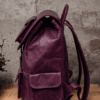 An aged purple leather backpack with multiple pockets on a wooden surface in front of a concrete wall.