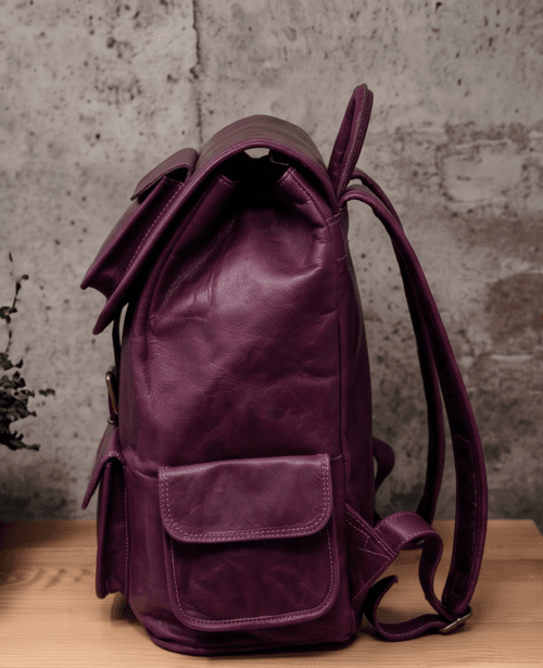 An aged purple leather backpack with multiple pockets on a wooden surface in front of a concrete wall.