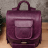 An aged purple leather backpack with multiple pockets on a wooden surface in front of a concrete wall.