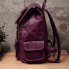 An aged purple leather backpack with multiple pockets on a wooden surface in front of a concrete wall.