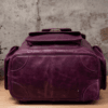 An aged purple leather backpack with multiple pockets on a wooden surface in front of a concrete wall.