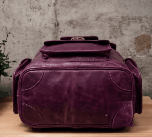 An aged purple leather backpack with multiple pockets on a wooden surface in front of a concrete wall.