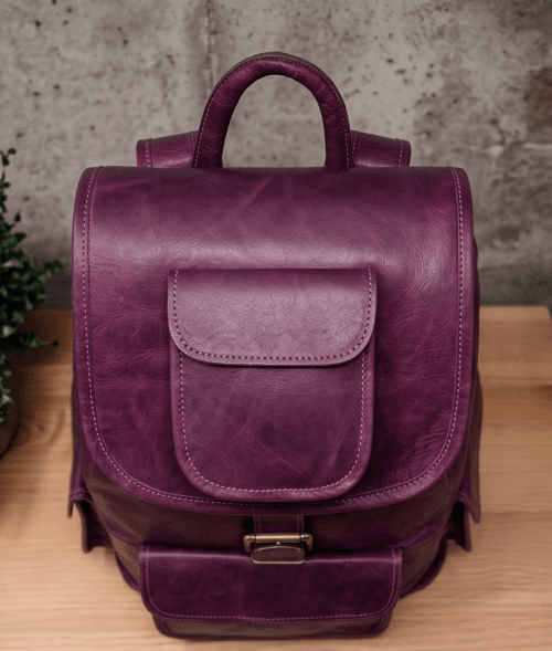 An aged purple leather backpack with multiple pockets on a wooden surface in front of a concrete wall.